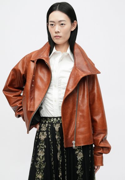 Smeđa kožna jakna s širokim okovratnikom, zatvaranjem na zip, i oversized krojem, uparena s bijelom košuljom i cvjetnom crnom suknjom.