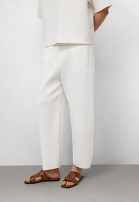 Weiße, weit geschnittene Hosen aus weichem Stoff, kombiniert mit braunen, schlichten Riemchensandalen. Glatte Textur, minimalistisches Design, keine sichtbaren Muster.