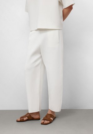 Pantalons blancs à jambes larges en tissu doux, associés à des sandales à lanières brunes. Texture lisse, design minimaliste, pas de motifs visibles.