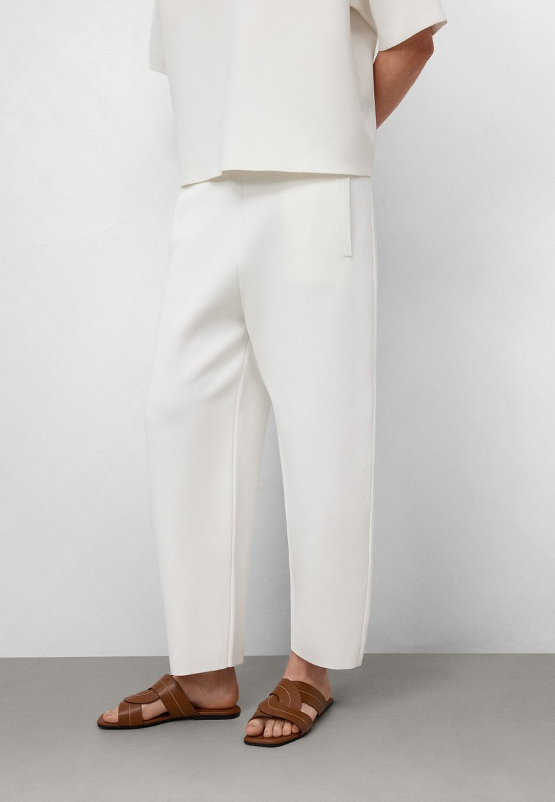Weiße, weit geschnittene Hosen aus weichem Stoff, kombiniert mit braunen, schlichten Riemchensandalen. Glatte Textur, minimalistisches Design, keine sichtbaren Muster.