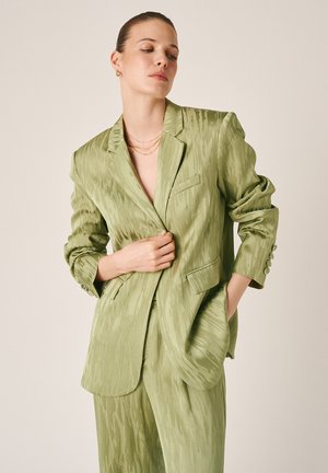 Donna che indossa un completo verde chiaro con blazer e pantaloni abbinati, in piedi con una mano che sistema il blazer e l'altra in tasca.