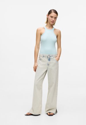 Donna in piedi con una mano in tasca, indossa un top senza maniche azzurro chiaro, jeans a gamba larga di denim chiaro e sandali neri.