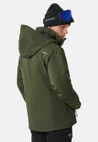 Grüne Skijacke mit Kapuze, Frontreißverschluss und Logodetail. Strukturiertes Finish und verstellbare Bündchen. Getragen mit einer schwarzen Mütze und Skibrille.