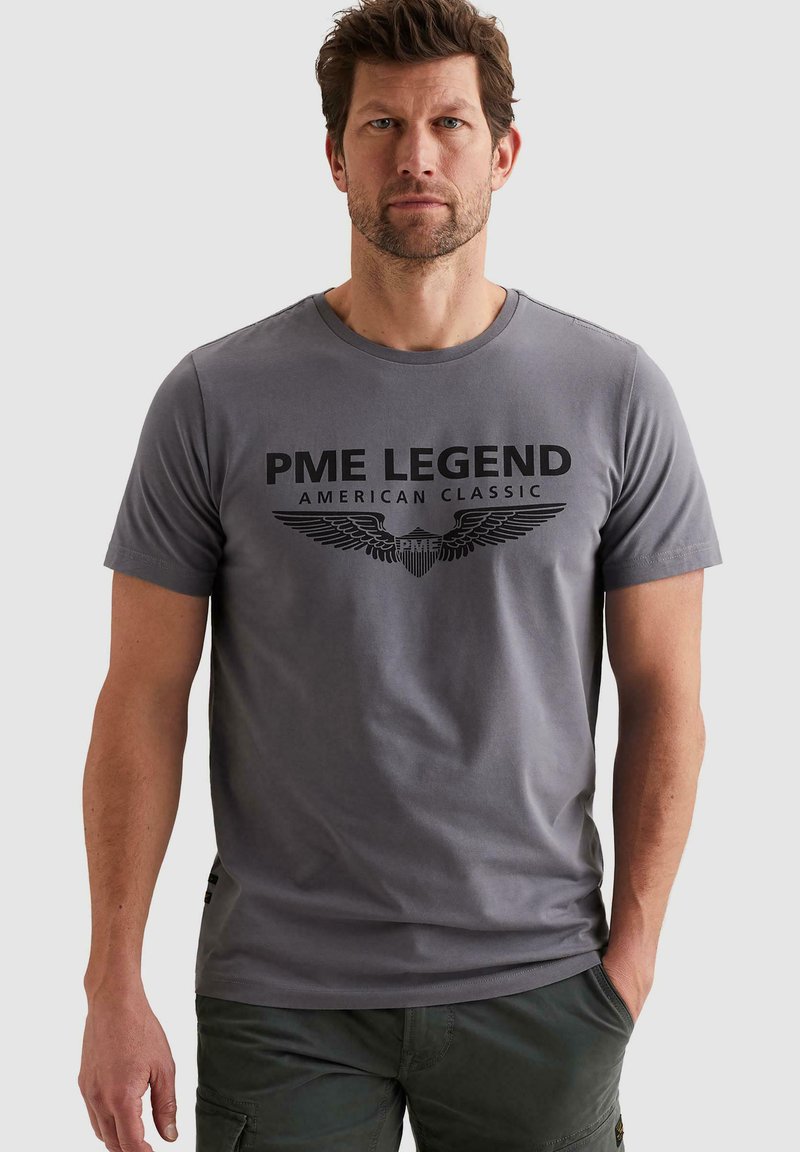 Homme aux cheveux courts bruns et barbe, portant un t-shirt gris PME Legend American Classic et un pantalon foncé, debout avec une main dans la poche.