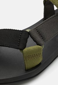 Sandalias de tiras negras y verde oliva con suela texturizada negra, que cuentan con tiras de nailon trenzadas y un detalle de conector de plástico.