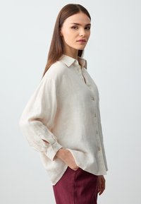 Camicia di lino color beige chiaro con colletto, chiusura a bottoni e maniche larghe. Presenta spacchi laterali e un tessuto morbido e texturizzato. Abbinata a pantaloni color bordeaux.