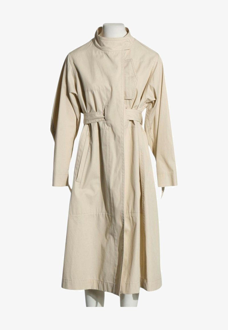 Trench-coat long beige avec col haut, manches longues, taille ceinturée, poches latérales et fermeture asymétrique devant sur un mannequin.