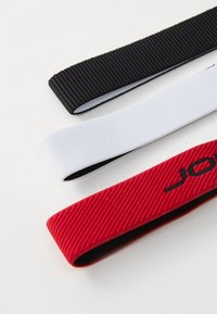 Tri elastične trake za kosu: crna s rebrastom teksturom, bijela s glatkom površinom i crvena s rebrastom teksturom koja ima crni logotip.