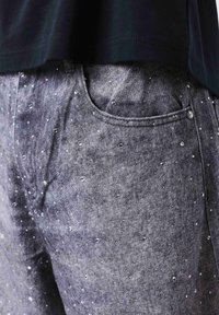 Gros plan d'un pantalon en jean gris avec de petits clous décoratifs argentés et une poche avant, porté avec un haut noir.