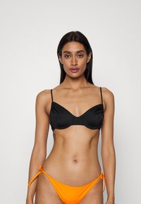 Top de biquíni preto com aros e alças finas, combinado com parte de baixo de biquíni laranja com laço lateral. Textura de tecido suave.