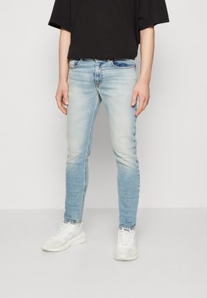 Vaqueros de mezclilla azul claro con un corte ajustado, lavado deslavado, cinco bolsillos y remaches de latón. Combinados con zapatillas blancas y una camiseta negra.