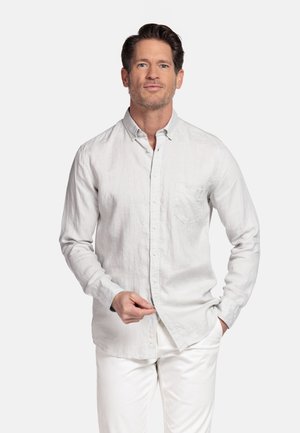 Man met kort donker haar, gekleed in een wit overhemd met knopen en witte broek, staand met één hand in de zak en de andere terwijl hij zijn overhemd rechtzet.