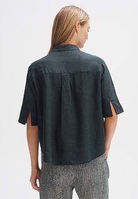 Maglietta a maniche corte in lino verde scuro con colletto, caratterizzata da spacchi laterali e una vestibilità rilassata. Dettaglio della cucitura posteriore visibile. Indossata con pantaloni a fantasia.