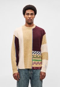 Kootud kampsun, millel on beežide, burgundia ja kreemikate toonidega patchwork-muster, tekstuursete mustrite ja siniste õmblusdetailidega. Ümmargune kaelus, lõdvestunud istuvus.