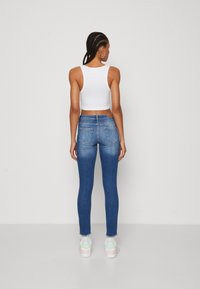 Žena s opletenými vlasmi, oblečená v bielom crop top, modrých úzkych džínsach, bielych ponožkách a bielych teniskách, stojí chrbtom k fotoaparátu na jednofarebnom pozadí.