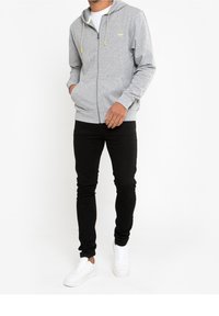 Gray zip-up hoodie with a hood, featuring yellow drawstrings and small logo. Paired with black skinny jeans and white sneakers.