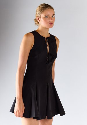 Jeune femme aux cheveux blonds portant une robe noire sans manches avec des liens à l'avant, debout devant un fond uni gris clair.