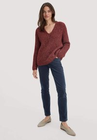 Weinroter gerippter Strickpullover mit tiefem V-Ausschnitt, kombiniert mit navyblauen Cordhosen und hellgrauen flachen Schuhen. Texturierte Stoffdetails sichtbar.