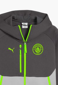 Šedá mikina na zip s neonově zelenými akcenty. Má kapuci, boční kapsy a emblém Manchester City na hrudi. Vyrobena z měkkého materiálu.