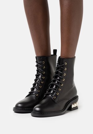 Botas de tobillo de cuero negro con ojales de metal dorado, diseño de cordones y un exclusivo tacón de bloque adornado con perlas. Textura suave, forma clásica.