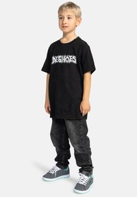 Černé tričko s bílým grafickým textem, spárované s tmavými denimovými džínami. Obuv je šedá s tyrkysovými akcenty a bílými tkaničkami. Dítě stojí.