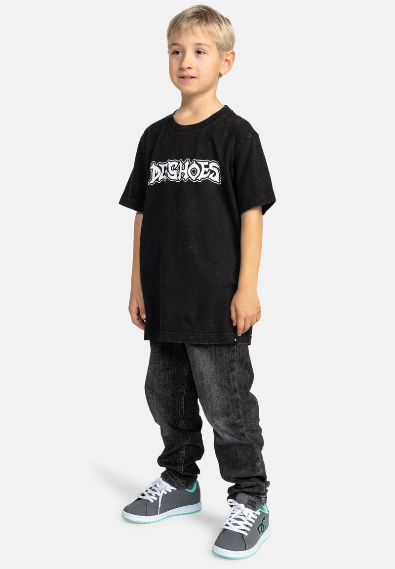 Černé tričko s bílým grafickým textem, spárované s tmavými denimovými džínami. Obuv je šedá s tyrkysovými akcenty a bílými tkaničkami. Dítě stojí.