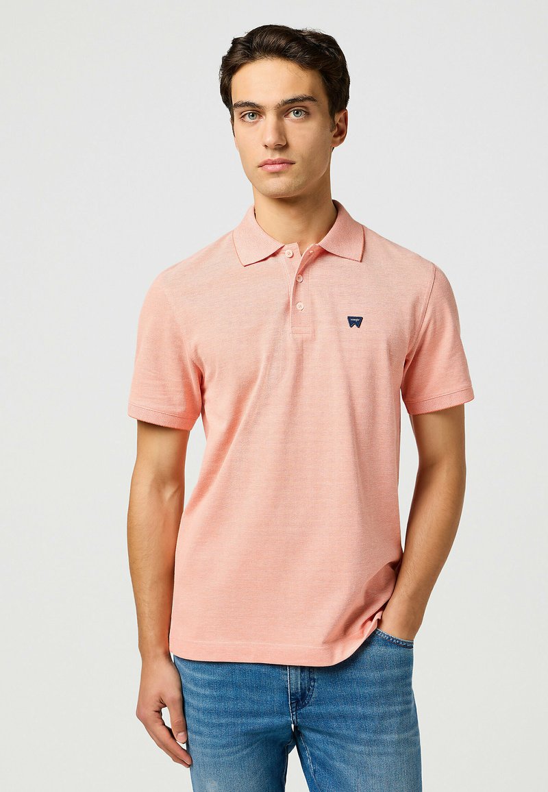 Polo in rosa chiaro, maniche corte, tessuto testurizzato, colletto con tre bottoni, piccolo logo scuro sul petto, abbinato a jeans blu.
