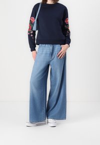 Persona vestida con un suéter azul marino con bordados florales rosas en las mangas, jeans de pierna ancha de color azul claro, zapatillas blancas y llevando un bolso de hombro azul.