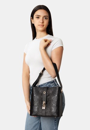 Young woman in white t-shirt holding black embossed leather handbag with shoulder strap and gold clasp, standing against plain background.