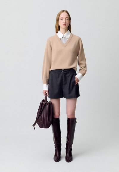 Pull à col en V beige, short gris cintré avec fermeture à double boucle, bottes en cuir bordeaux arrivant au genou et un sac seau assorti.