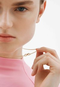 Collarín de oro con el símbolo del infinito sobre una delicada cadena, sostenido por una mano, con un fondo blanco liso. La modelo lleva una blusa rosa.