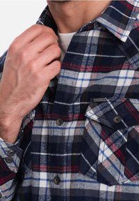 Koszula flanelowa w kratę w kolorze granatowym, białym i czerwonym. Posiada kołnierzyk zapinany na guziki, dwie kieszenie na piersi oraz czarne guziki przy zapięciu z przodu.