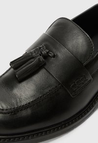 Mocassins en cuir noir avec une finition lisse, une sangle décorative avec des franges et des détails de couture le long de la semelle.