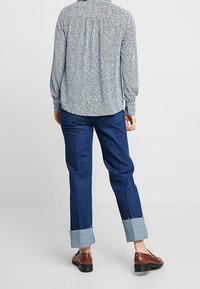 Weißes und marineblau gemustertes Langarmhemd mit gerafftem Rücken, kombiniert mit einer dunklen Jeans mit umgeschlagenen Säumen und braunen Schuhen.