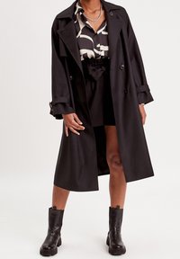 Sort trenchcoat med dobbeltrækkede knapper, brede revers og sidelommer, lagdelt over en mønstret skjorte og sorte shorts. Sorte ankelstøvler.