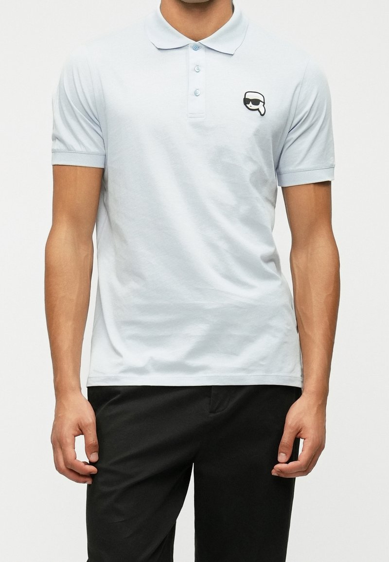 Homme portant un polo bleu clair à trois boutons et un pantalon noir, debout les bras détendus le long du corps.
