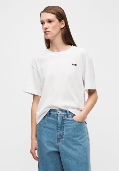 Άσπρο βαμβακερό t-shirt με κοντά μανίκια και στρογγυλή λαιμόκοψη, με μικρή μαύρη ετικέτα στην αριστερή πλευρά. Συνδυασμένο με ανοιχτό μπλε τζιν παντελόνι.