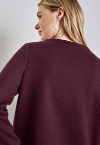 Burgundy teksturoitu neulepusero, jossa on geometrinen kuvio, pyöreä kaula-aukko ja pitkät hihat. Sujuva kangas laskeutuu hartioiden yli.