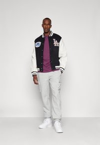 Varsityjack in zwart en wit, met LA-embleem, gecombineerd met een paarse t-shirt, grijze sweatpants en witte sneakers.