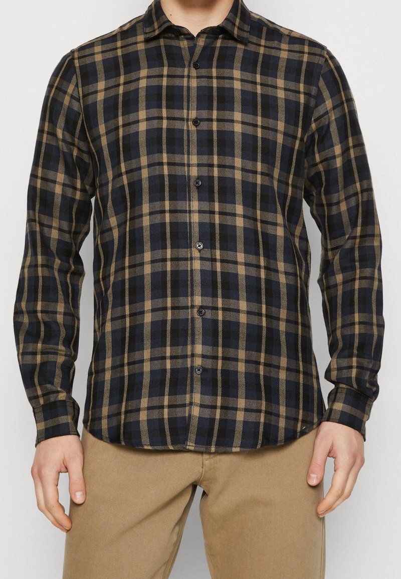 Homme portant une chemise à manches longues avec un motif à carreaux noir et marron et un pantalon marron clair sur un fond uni.