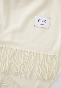 Cremfarvet cashmere tørklæde med frynser og et hvidt mærke med teksten "FTC Cashmere" syet på den ene side.