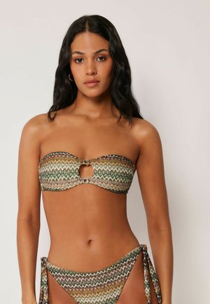 Kobieta ubrana w wzorzysty bikini bez ramiączek z wycięciem z przodu i wiązaniami po bokach, stojąca na tle jednolitego, jasnego tła.