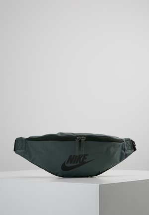 Dunkelgrüne Nike-Gürteltasche mit schwarzem Logo und Doppelreißverschluss, auf einem weißen Sockel vor einem schlichten grauen Hintergrund präsentiert.