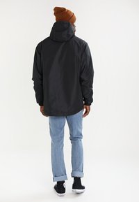 Mężczyzna w czarnej kurtce z kapturem, brązowej czapce beanie, jasnoniebieskich jeansach z mankietami i czarnych sneakersach na wysokiej podeszwie stoi tyłem.