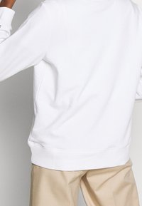 En person som bär beige byxor och en enkel vit långärmad tröja, sedd bakifrån mot en neutral bakgrund.
