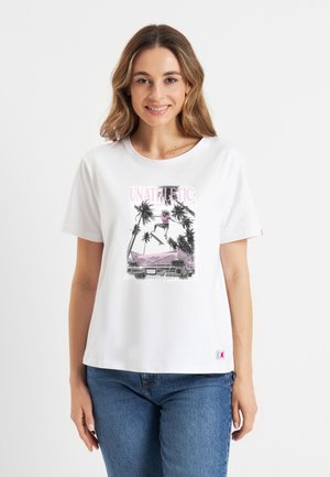 Lachende vrouw die een wit T-shirt draagt met een afbeelding van een skateboarder die over een roze auto springt, omgeven door palmbomen en het woord "UNATHLETIC."