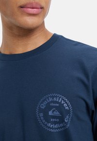 Camiseta azul marino con cuello redondo y mangas cortas, que presenta un estampado gráfico circular gris de "Quiksilver" y un logo de montaña.