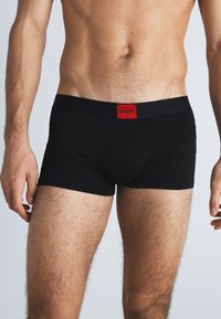 Schwarze Baumwoll-Boxershorts mit glatter Textur, ausgestattet mit einem roten, gebrandeten Bund. Das Design ist figurbetont und minimalistisch.