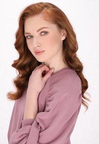 Felpa di color rosa chiaro, con texture e dettagli delle maniche arricciati, indossata da una donna con capelli rossi ondulati, in posa contro uno sfondo bianco.