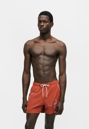 Athletischer Mann trägt orangefarbene Badehose mit weißem Kordelzug und abstrakten Linienmustern, steht vor einem schlichten hellen Hintergrund.